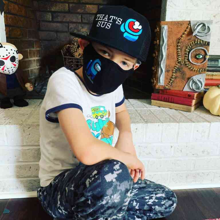 A young boy sitting on the floor with a mask on his face, wearing That's Sus Hat.
