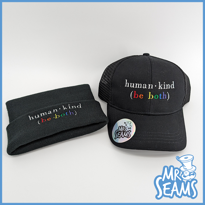 Two black headwear items, one beanie and one baseball cap from the Human - Kind (Be Both) Hats & Beanies collection, displayed against a light background.