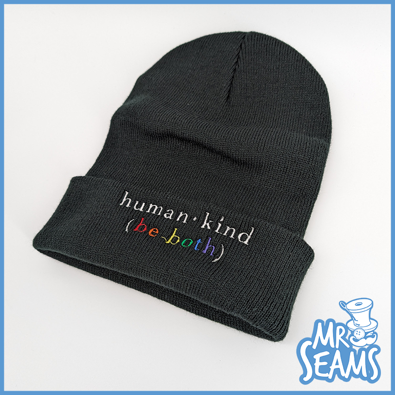 A black beanie from the Human - Kind (Be Both) Hats & Beanies collection, featuring the stitched phrase, "human.kind (be both)," showcased on a white background.