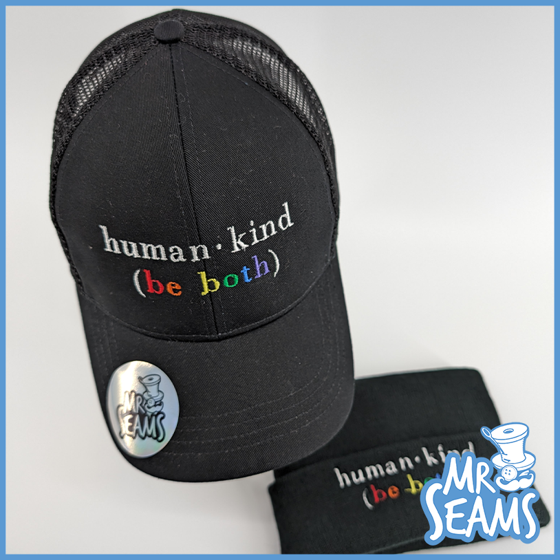 A black Human - Kind (Be Both) hat with the phrase "Human.Kind (Be Both)" embroidered on it, accompanied by a pin and a cloth wristband with the same phrase.